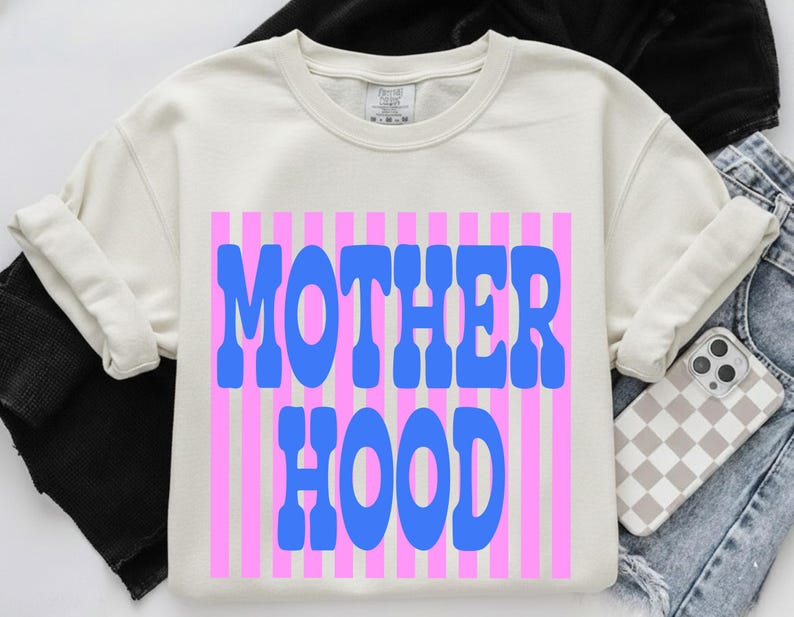 May include: A cream-colored crewneck sweatshirt featuring a graphic print of vertical pink stripes with the word MOTHER HOOD written in large, bold, blue letters across the center. The garment has a ribbed neckline and cuffed sleeves.