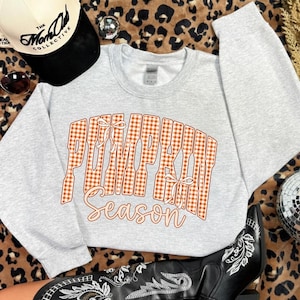 May include: A light gray crewneck sweatshirt featuring the text PUMPKIN SEASON in an orange and white gingham pattern with two small white bows integrated into the lettering. The sweatshirt is displayed alongside a baseball cap and a boot.