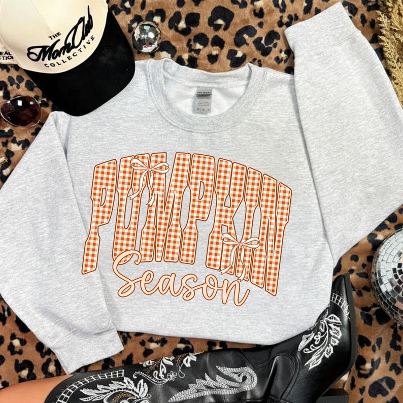 May include: A light gray crewneck sweatshirt featuring the text PUMPKIN SEASON in an orange and white gingham pattern with two small white bows integrated into the lettering. The sweatshirt is displayed alongside a baseball cap and a boot.