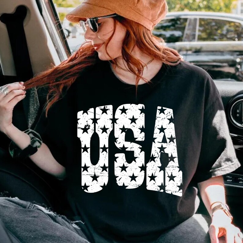 May include: Black t-shirt with the word "USA" in large, distressed white letters filled with black stars. The shirt is a classic crew neck style.
