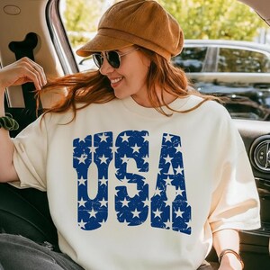 May include: Cream-colored sweatshirt with the word "USA" in distressed blue lettering filled with white stars. The person in the image is wearing a brown corduroy cap and sunglasses. The overall theme is patriotic.
