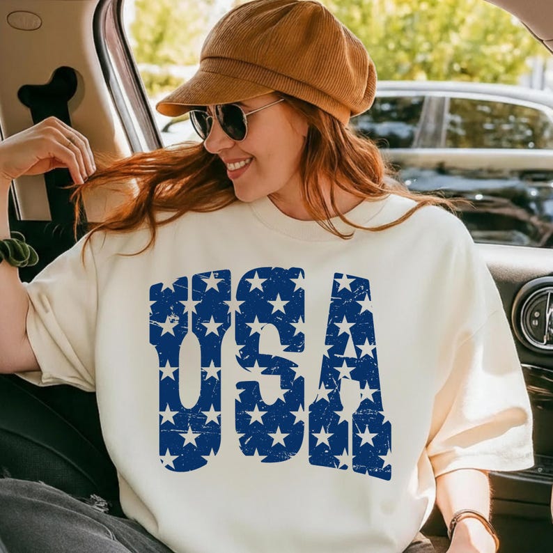 May include: Cream-colored sweatshirt with the word "USA" in distressed blue lettering filled with white stars. The person in the image is wearing a brown corduroy cap and sunglasses. The overall theme is patriotic.