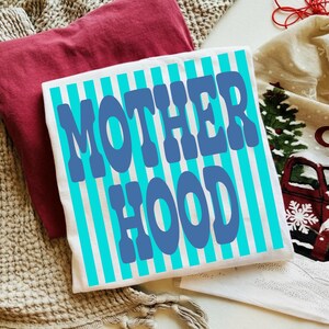 May include: A white t-shirt featuring vertical cyan stripes and the text MOTHER HOOD printed in large, bold blue letters across the center. The shirt is displayed alongside a folded maroon garment and a cream-colored fabric with a red truck print.