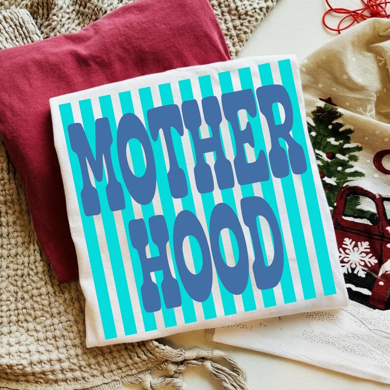 May include: A white t-shirt featuring vertical cyan stripes and the text MOTHER HOOD printed in large, bold blue letters across the center. The shirt is displayed alongside a folded maroon garment and a cream-colored fabric with a red truck print.