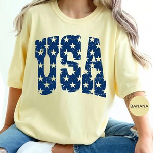 May include: A light yellow t-shirt with the word "USA" in distressed navy blue lettering filled with white stars. The shirt has short sleeves and a round neckline. The word "BANANA" is printed on a small, round sticker on the arm.