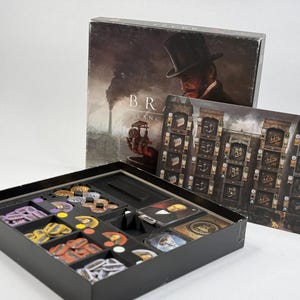 Könnte beinhalten: Ein Brettspiel-Set mit dem Titel Brass: Lancashire, bestehend aus einem Schachteldeckel mit einem Mann im Zylinder, einem Spielplan und einem schwarzen Kunststoffeinsatz mit verschiedenen runden Spielsteinen, Karten und Kleinteilen.