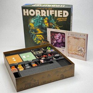 Op de afbeelding: Het bordspel Horrified American Monsters bevat een grote geïllustreerde doos, een spelhandleiding met een skelet-violist op de omslag en een zwart plastic inzetbakje met diverse gekleurde spelfiches en speelkaarten.