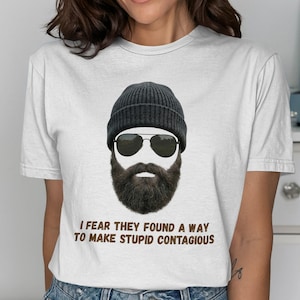 May include: White t-shirt featuring a graphic of a bearded face wearing a black beanie and sunglasses. The text "I fear they found a way to make stupid contagious" is printed below the graphic in brown.