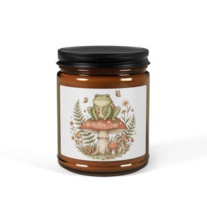 May include: A brown glass jar with a black lid. The jar has a white label with a whimsical illustration of a frog wearing a vest sitting on a mushroom. The illustration includes ferns, flowers, and butterflies.