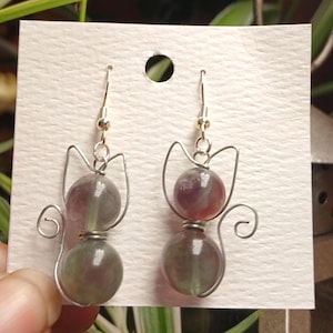 Pode incluir: Um par de brincos pendentes com um design de silhueta de gato feito de arame. Cada brinco consiste em duas contas de pedra redondas, roxas e verdes, empilhadas verticalmente, com arame prateado formando as orelhas e uma cauda enrolada.
