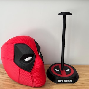 May include: A red and black Deadpool mask and stand. The mask features a textured surface and black eye cutouts. The stand is black with a red Deadpool logo and the word "DEADPOOL" on the base.