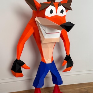 Op de afbeelding: Een grote, geometrische papieren sculptuur van Crash Bandicoot. De figuur is voornamelijk oranje met bruine oren, zwarte neus en handen, een blauwe broek en rode schoenen met witte strepen. De sculptuur wordt tentoongesteld tegen een witte muur en een houten vloer.