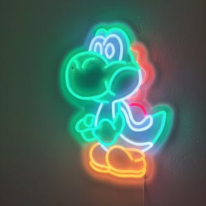 Pu&ograve; includere: Insegna al neon a forma di personaggio di dinosauro dei cartoni animati, con contorno verde e interno blu. Il dinosauro ha i piedi arancioni e un accento rosso sulla schiena. L'insegna &egrave; collegata alla parete.