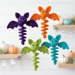 May include: Four hanging crochet bat decorations in purple, orange, lime green, and teal. Each bat features a round head with pointed ears, two outstretched wings, and a long, spiralling tail hanging downward, crafted from yarn with visible stitches.