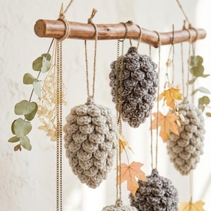 May include: A wall hanging featuring a horizontal wooden branch suspended by twine. Hanging from the branch are several crocheted pinecones in shades of beige and grey, interspersed with dried eucalyptus sprigs, gold chains, and orange maple leaves.
