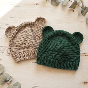 May include: Two handmade crochet beanies featuring rounded bear ears on top. One beanie is a light tan color, and the other is a deep forest green. Both hats have a ribbed brim and a textured stitch pattern throughout the main body of the headwear.