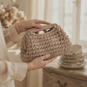 Peut inclure: Un sac à main en crochet beige présentant un motif en maille épaisse et texturée avec une poignée intégrée. Le sac a une forme rectangulaire structurée avec un design de points visible sur tout l'extérieur, fait d'un matériau épais.