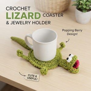 May include: A green crochet lizard coaster and jewelry holder with a white mug placed on top. The lizard features white eyes with black pupils and a bright pink berry in its open mouth. Text on the image reads CROCHET LIZARD COASTER & JEWELRY HOLDER, Popping Berry Design!, and CUTE & USEFUL!