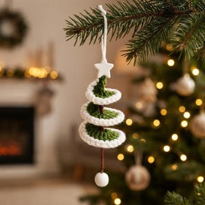 Puede incluir: Un adorno para árbol de Navidad en forma de espiral, hecho de hilo verde oscuro con bordes tejidos en blanco. El adorno presenta una estrella blanca en la parte superior, un eje central de hilo marrón y una pequeña cuenta de madera blanca.