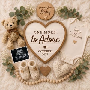 May include: A beige crochet teddy bear with a brown bow, a heart-shaped letter board reading ONE MORE to Adore OCTOBER 2027, a cream baby bodysuit labeled Baby Williams, an ultrasound photo, cream booties, a wooden sign, and greenery.