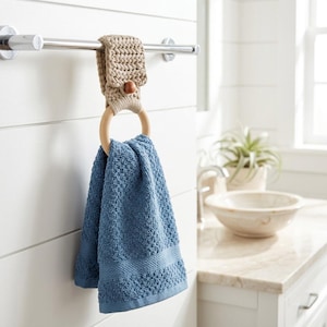 May include: A blue textured hand towel hangs from a light wood ring, which is attached to a chrome towel bar by a beige crocheted fabric strap secured with a small brown wooden button. The towel features a woven border detail at the bottom.