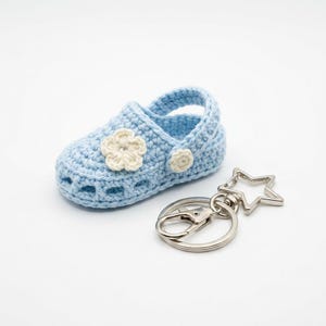 May include: A light blue crochet baby shoe with a cream-colored flower accent. A silver-colored star-shaped keychain is attached. The shoe is approximately 3 inches long. The item is set against a white background.