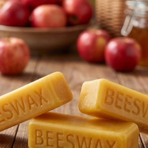 May include: Close-up of three rectangular bars of beeswax with the word "BEESWAX" embossed on them. The bars are a warm yellow color and are arranged on a wooden surface. Red apples and a basket are in the background.