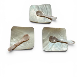 May include: Three square mother-of-pearl dishes with matching small spoons. The dishes and spoons have a pearlescent sheen with subtle variations in color. The spoons are resting on the dishes, creating a set.
