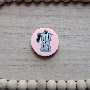 May include: A circular pink pin featuring a silver and black illustration of a moka pot coffee maker. The design is centered on the disc, showcasing the classic octagonal shape, handle, and spout of the stovetop espresso brewer in a flat graphic style.