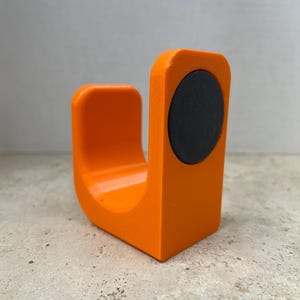 Peut inclure: Objet orange vif en forme de U avec un aimant circulaire noir fix&eacute; sur un c&ocirc;t&eacute;. L'objet, probablement imprim&eacute; en 3D, repose sur une surface textur&eacute;e. Le design est simple et moderne, avec des bords arrondis.