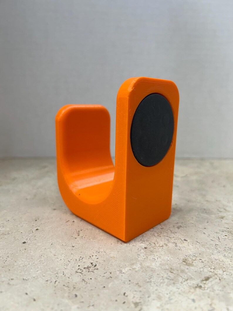 Peut inclure: Objet orange vif en forme de U avec un aimant circulaire noir fix&eacute; sur un c&ocirc;t&eacute;. L'objet, probablement imprim&eacute; en 3D, repose sur une surface textur&eacute;e. Le design est simple et moderne, avec des bords arrondis.