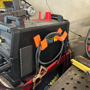 Peut inclure: Un poste &agrave; souder noir et orange avec le mot "new" visible. La machine a une poign&eacute;e et est connect&eacute;e &agrave; un c&acirc;ble noir maintenu par des clips orange. Une pince dor&eacute;e est attach&eacute;e au c&acirc;ble. Une structure de support rouge est visible.