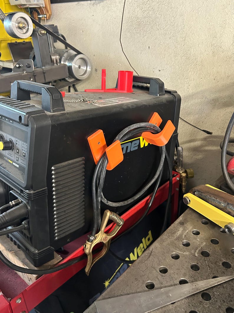 Peut inclure: Un poste &agrave; souder noir et orange avec le mot "new" visible. La machine a une poign&eacute;e et est connect&eacute;e &agrave; un c&acirc;ble noir maintenu par des clips orange. Une pince dor&eacute;e est attach&eacute;e au c&acirc;ble. Une structure de support rouge est visible.