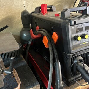 Peut inclure: Un poste &agrave; souder PrimeWeld MIG200 noir et rouge avec des supports de c&acirc;bles orange et rouges. Le poste &agrave; souder a une poign&eacute;e noire et des boutons jaunes. Une bouteille de gaz grise est visible &agrave; gauche.