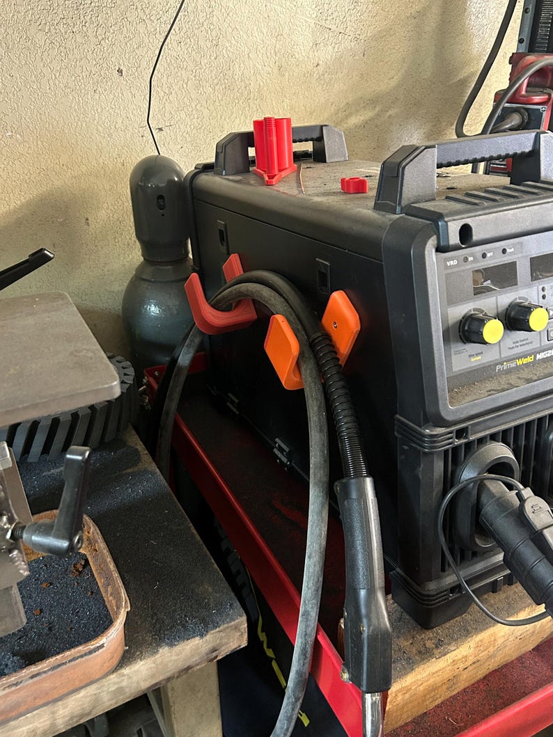 Peut inclure: Un poste &agrave; souder PrimeWeld MIG200 noir et rouge avec des supports de c&acirc;bles orange et rouges. Le poste &agrave; souder a une poign&eacute;e noire et des boutons jaunes. Une bouteille de gaz grise est visible &agrave; gauche.