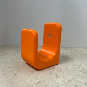 Peut inclure: Support en forme de U de couleur orange vif, avec une base incurv&eacute;e et deux c&ocirc;t&eacute;s verticaux. Une seule vis est visible sur un c&ocirc;t&eacute;. Le support est fait d'un mat&eacute;riau lisse et solide et repose sur une surface textur&eacute;e.