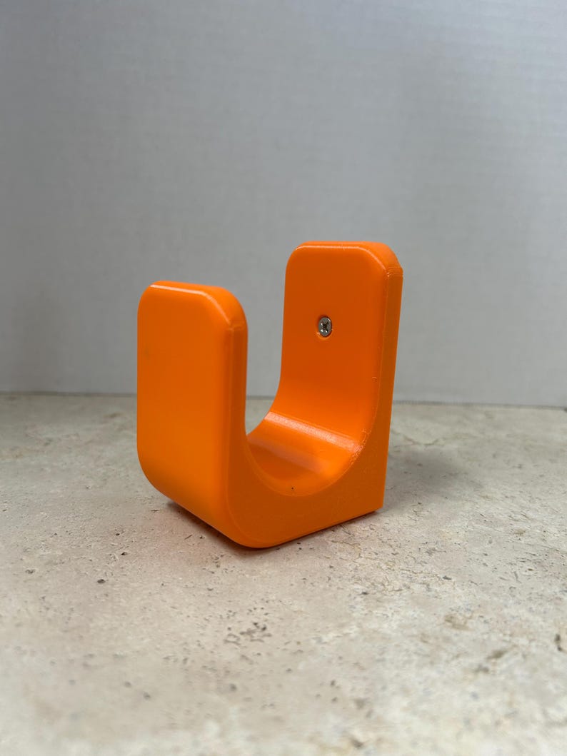 Peut inclure: Support en forme de U de couleur orange vif, avec une base incurv&eacute;e et deux c&ocirc;t&eacute;s verticaux. Une seule vis est visible sur un c&ocirc;t&eacute;. Le support est fait d'un mat&eacute;riau lisse et solide et repose sur une surface textur&eacute;e.