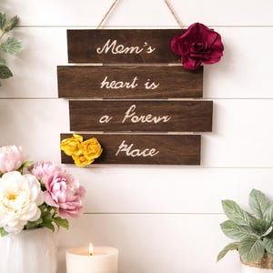 May include: A wooden sign with the words "Mom's heart is a forever place" in white script. The sign is decorated with a burgundy and yellow flower. A vase of flowers and a lit candle are in the foreground.