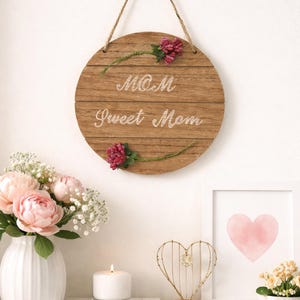 May include: A round wooden sign with the words "MOM Sweet Mom" in white script, adorned with pink floral accents. The sign hangs from a jute rope. A vase of pink flowers and a framed heart are also visible.