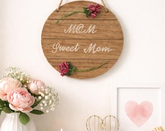 Può includere: Un cartello rotondo in legno con le parole "MOM Sweet Mom" in scrittura bianca, adornato con decorazioni floreali rosa. Il cartello è appeso a una corda di iuta. Sono visibili anche un vaso di fiori rosa e un cuore incorniciato.
