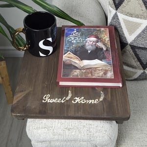 May include: A dark brown wooden sofa table with the text "Sweet Home" and a decorative wreath. A black mug with a gold handle and the letter "S" sits on the table, along with a book. A houseplant and a patterned pillow are also visible.