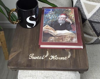 May include: A dark brown wooden sofa table with the text "Sweet Home" and a decorative wreath. A black mug with a gold handle and the letter "S" sits on the table, along with a book. A houseplant and a patterned pillow are also visible.