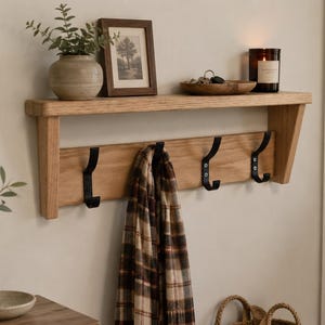 May include: A wooden wall shelf with a lower section featuring three black hooks. A framed picture, a small vase with greenery, and a candle sit on the shelf. A plaid scarf hangs from the hooks. The shelf is mounted on a light-colored wall.