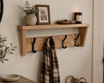 Può includere: Una mensola a muro in legno con una sezione inferiore con tre ganci neri. Una foto incorniciata, un piccolo vaso con verde e una candela sono sulla mensola. Una sciarpa a quadri è appesa ai ganci. La mensola è su una parete chiara.