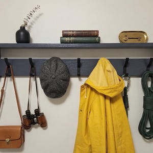 May include: A dark gray wooden wall-mounted shelf with five black hooks. Items hanging include a tan leather bag, binoculars, a gray tweed flat cap, a yellow hooded raincoat, and a green rope. The shelf holds a black vase, two books, and a gold tray.