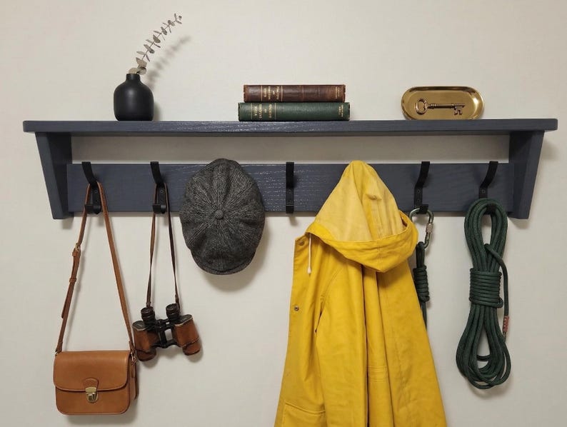 May include: A dark gray wooden wall-mounted shelf with five black hooks. Items hanging include a tan leather bag, binoculars, a gray tweed flat cap, a yellow hooded raincoat, and a green rope. The shelf holds a black vase, two books, and a gold tray.