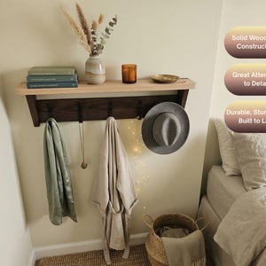 May include: A wall-mounted shelf with a light wood top and a dark wood base featuring four black metal hooks. The shelf holds books, a vase with dried plants, a candle, and a small dish. Text on the image reads: Solid Wood Oak Construction, Great Attention to Detail, Durable, Sturdy, and Built to Last.