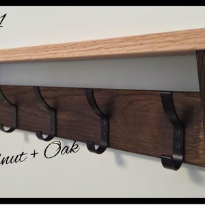 May include: A wall-mounted coat rack featuring a light-colored oak shelf atop a dark walnut wood backboard. The unit includes five black metal hooks and is labeled with the text Code 11 and D Walnut + Oak, along with a circular logo in the corner.