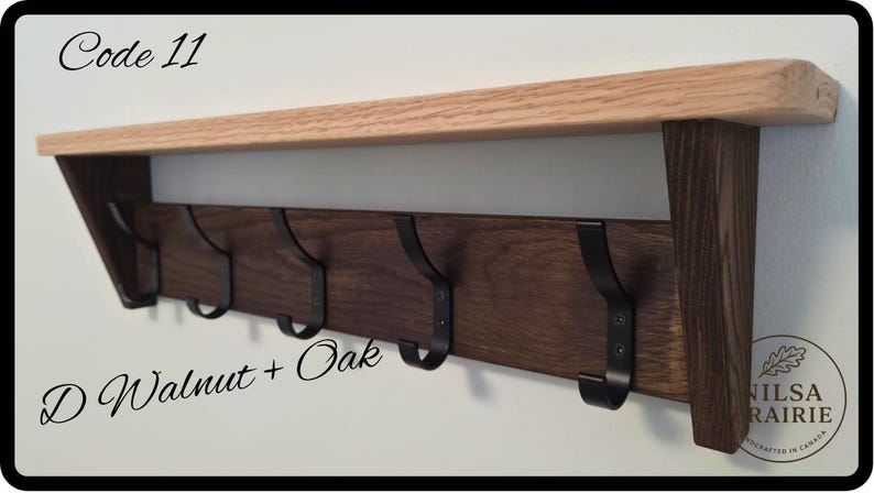 May include: A wall-mounted coat rack featuring a light-colored oak shelf atop a dark walnut wood backboard. The unit includes five black metal hooks and is labeled with the text Code 11 and D Walnut + Oak, along with a circular logo in the corner.