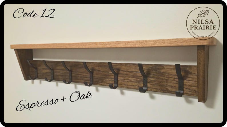 May include: A wall-mounted wooden shelf with a natural oak top and a dark espresso-stained backboard featuring seven black metal hooks. The text Code 12 and Espresso + Oak are visible, along with a circular logo reading NILSA PRAIRIE.